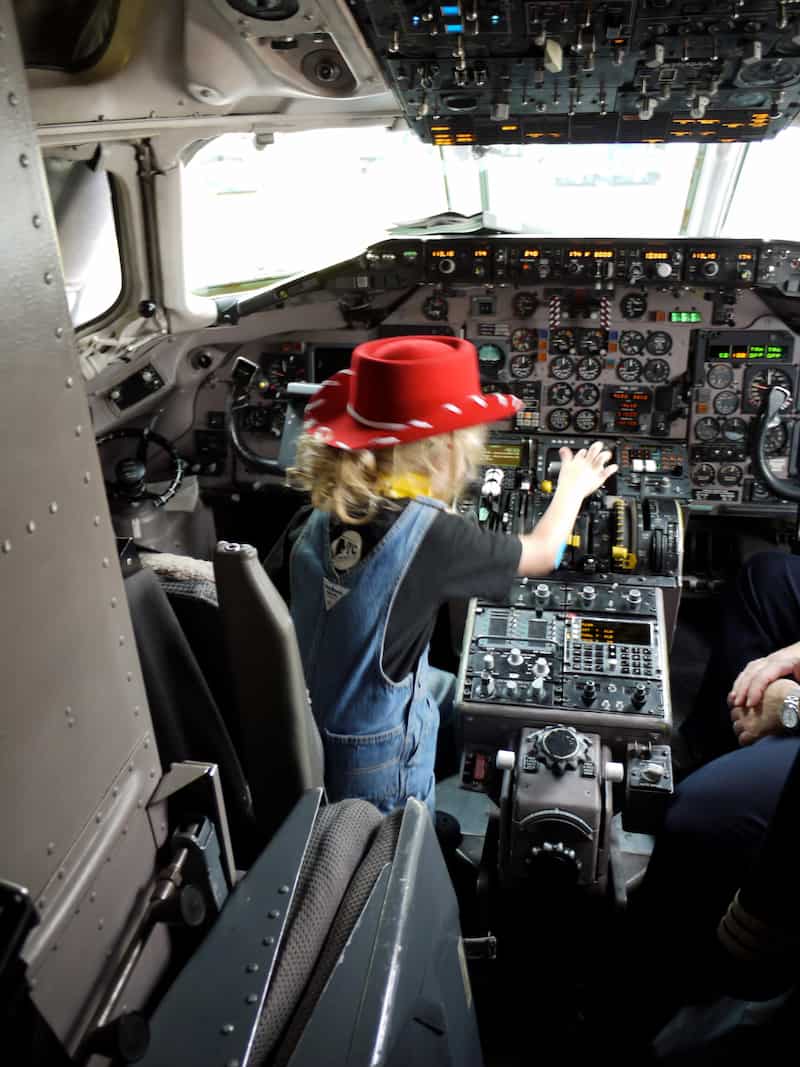 An airliner cockpit filled with analog gauges. Sitting in the pilot seat, a blurry child with a blond ponytail, red cowboy hat, and overalls.