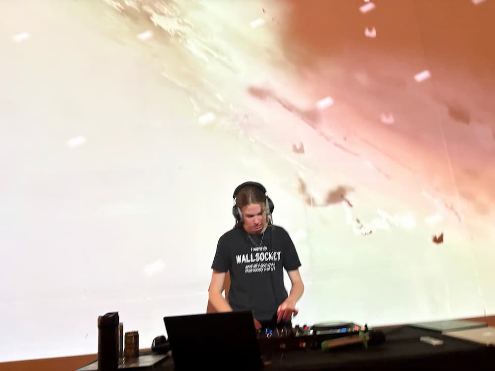 Me in a ponytail with bright visuals behind me focusing intently on the DJ controller on the table in front of me.