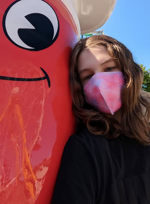 A light-skinned person with wavy shoulder-length brown hair hugging a red plastic human-sized Jelly Belly mascot man.