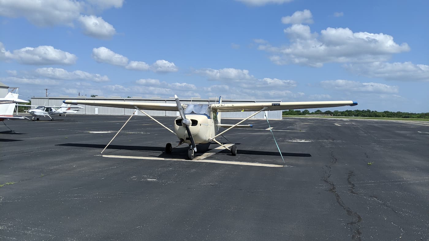 A small Cessna plane sitting in a parking area, taken from the front. A beautiful blue sky with spread out fluffy clouds is on top.
