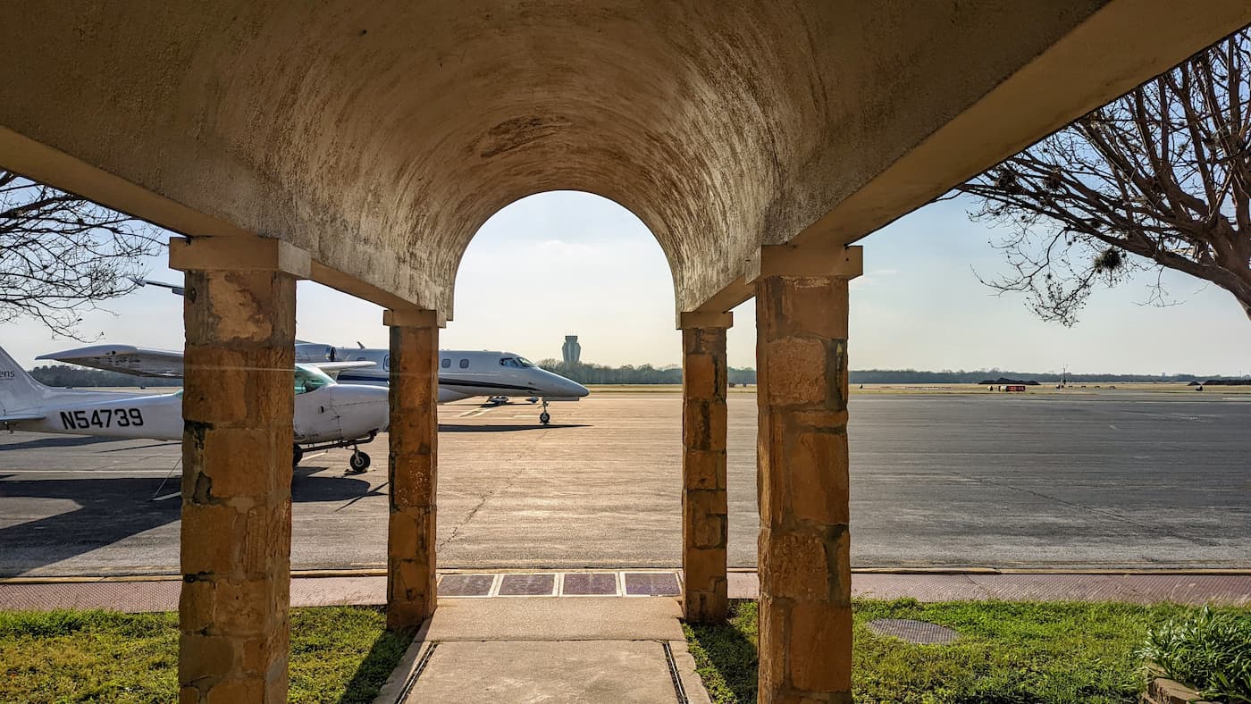 The parking area of a small airport, with two visible small planes. In the background is the runway and air traffic conctrol. The photograph is taken from the perspective of standing at the door, and is partially obscured by a brown ceiling held up by columns.