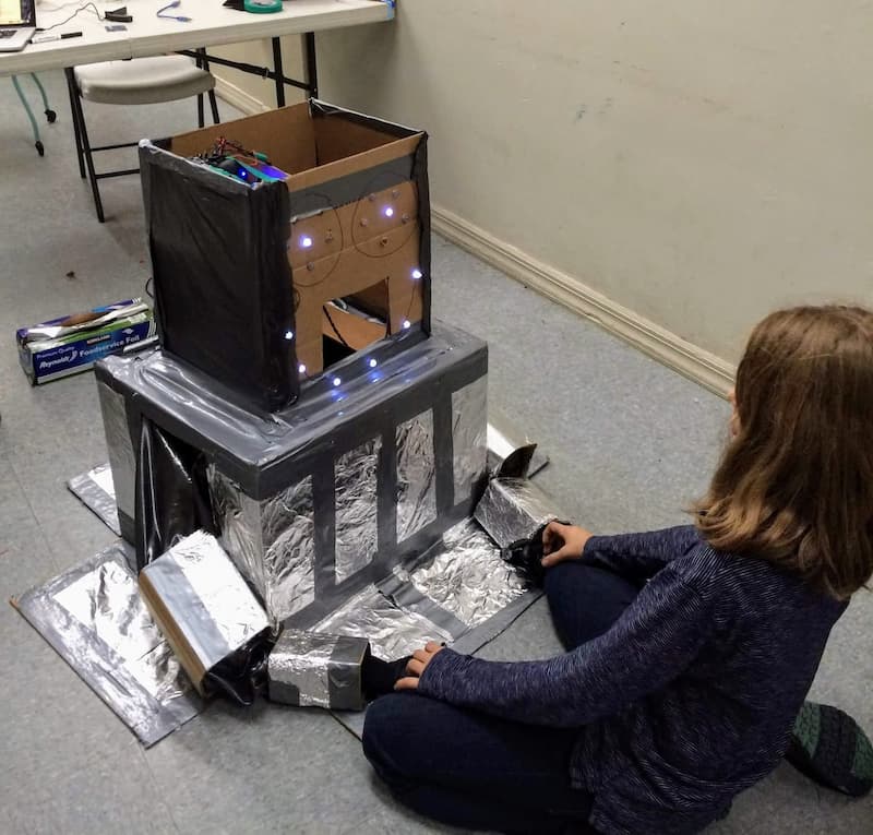 A young girl holding hands with a robot made mostly out of cardboard and covered in tinfoil. The face of the robot has lit LEDs and some electronics can be seen inside. A desk with wires and a roll of tinfoil can be seen in the background.