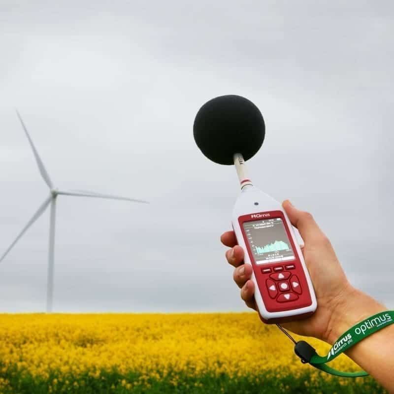 A sound level meter held out to measure the volume of a windmill in a field.